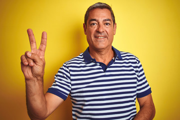 Handsome middle age man wearing striped polo standing over isolated yellow background smiling with happy face winking at the camera doing victory sign. Number two.