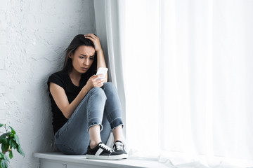 depressed young woman sitting on window sill at home and using smartphone - obrazy, fototapety, plakaty