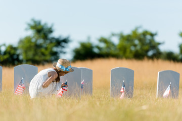 cute kid sitting near headstone with american flag in graveyard
