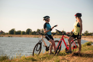 Fototapeta premium Two women cyclists with bikes standing and talking at the lake shore