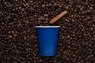 A blue disposable cup of coffee to go with a cinnamon stick against the background of roasted beans.
