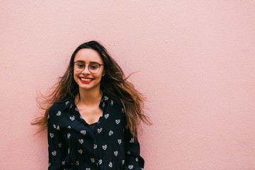 Charming teen woman in eyeglasses and blue shirt smiling happily at camera standing against pink wall