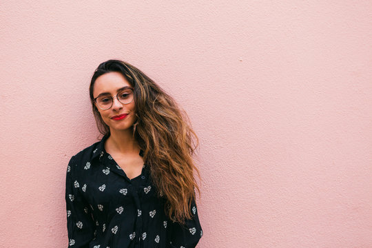 Charming teen woman in eyeglasses and blue shirt smiling happily at camera standing against pink wall