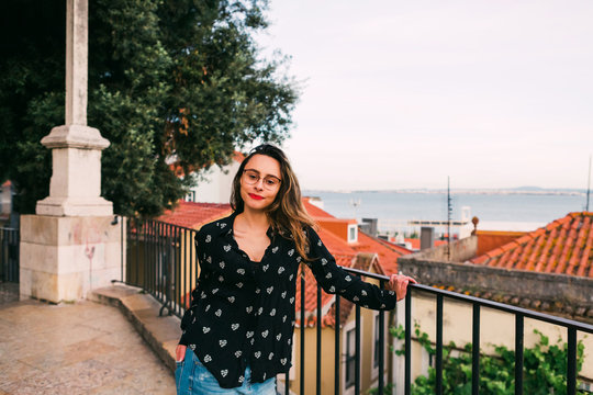 Portrait of smiling young brunette in glasses and with red lips looking at camera against blurred cityscape background