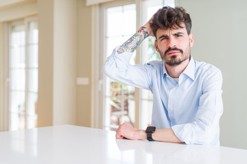 Young businesss man sitting on white table confuse and wonder about question. Uncertain with doubt, thinking with hand on head. Pensive concept.