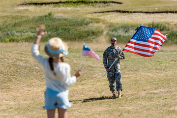 back view of kid in straw hat waving hand while holding american flag near man in military uniform