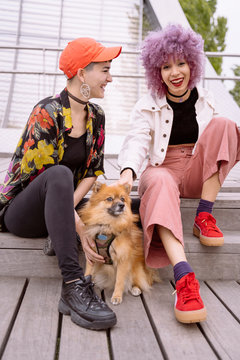 Light brown dog training by young attractive women with black and pink hair in bright clothes sitting on asphalt