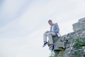 Concept: Working anytime and anywhere. Young handsome successful man is sitting on on the top of the mountain and working with digital tablet during summer journey.