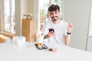Young man eating asian sushi from home delivery and ordering food using smartphone app very happy pointing with hand and finger to the side