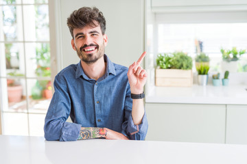Young man wearing casual shirt sitting on white table with a big smile on face, pointing with hand and finger to the side looking at the camera.