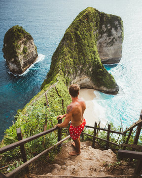From above of man standing on small terrace with fence observing picturesque ocean landscape with green cliffs on shore, Bali