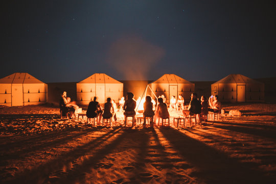 Group of travelers sitting around burning bright bonfire on sand in desert with tents, Morocco