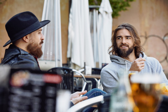 Handsome man in black hat sitting and enjoying conversation with friend wearing gray hoodie in outside cafe
