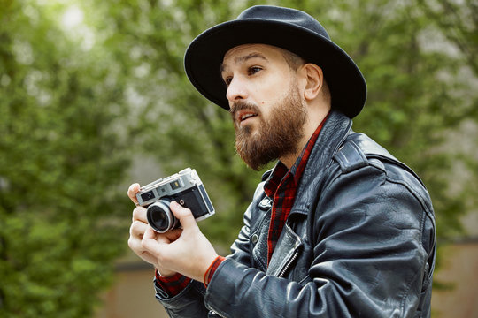 Young bearded handsome man in black hat and leather jacket taking photo in greenery street