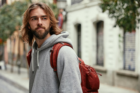 Young bearded handsome man in grey hoodie with backpack seriously walking outside