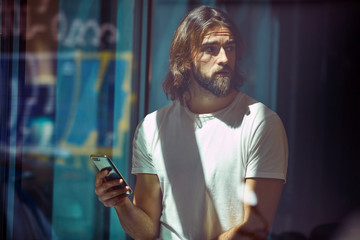 Young bearded handsome man leaning on wall with mobile phone in hands thoughtfully looking along
