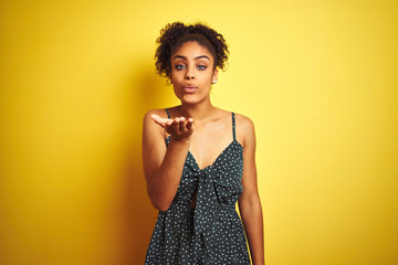African american woman wearing summer casual green dress over isolated yellow background looking at the camera blowing a kiss with hand on air being lovely and sexy. Love expression.