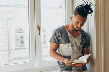 Young African American man with mohawk pigtails standing leaning on windowsill and reading book indoors