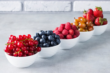 Fresh berries in white bowls on a gray background. Blueberries, strawberry, raspberry, gooseberries...