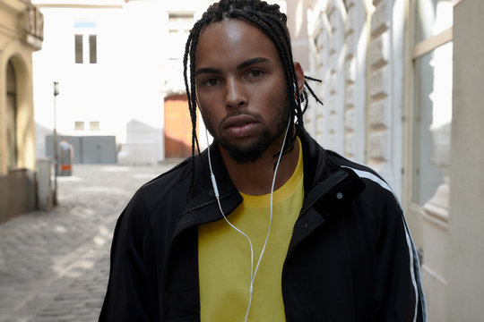 Serious young man with braids and earphones and mobile phone standing and looking at camera on urban background