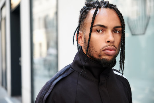 Young ethnic man with braided hair wearing black sports suit looking at camera on urban background