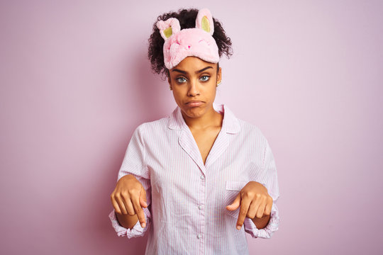 Young african american woman wearing pajama and mask over isolated pink background Pointing down looking sad and upset, indicating direction with fingers, unhappy and depressed. - Powered by Adobe