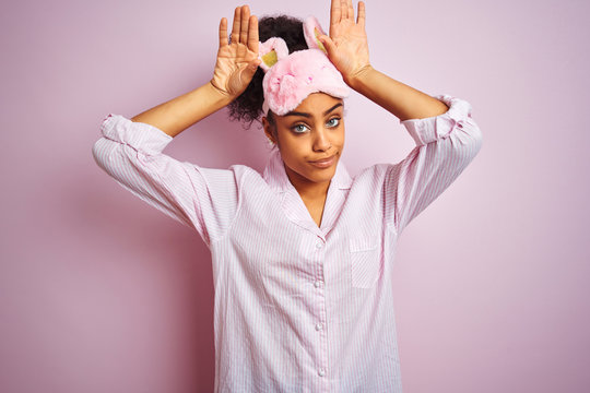Young african american woman wearing pajama and mask over isolated pink background Doing bunny ears gesture with hands palms looking cynical and skeptical. Easter rabbit concept. - Powered by Adobe