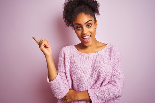 Young african american woman wearing winter sweater standing over isolated pink background with a big smile on face, pointing with hand finger to the side looking at the camera.