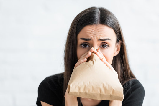 scared woman breathing into paper bag and looking at camera whule suffering from panick attact at home