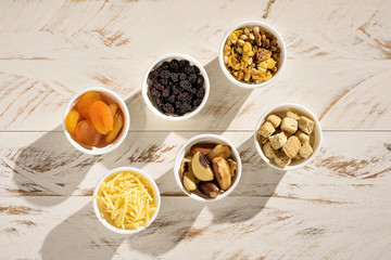 Six ramekins with cereal, croutons, brazil nuts, parmesan cheese, turkish apricot and raisin, on a white rustic wooden table