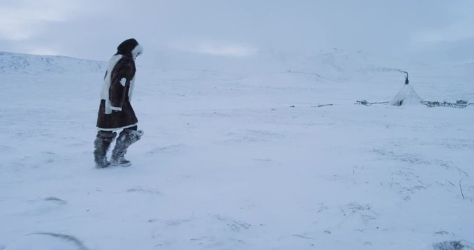 Closeup video of drone in the middle of Arctic walking man through the snowy field to the yurts he wearing traditional Yamal clothes from fur.