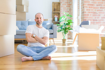 Fototapeta premium Young bald man sitting on the floor around cardboard boxes moving to a new home happy face smiling with crossed arms looking at the camera. Positive person.
