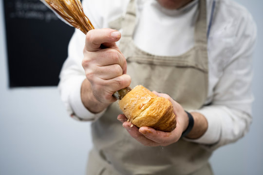 Crop man in apron and white uniform stuffing fresh croissant with chocolate cream indoors