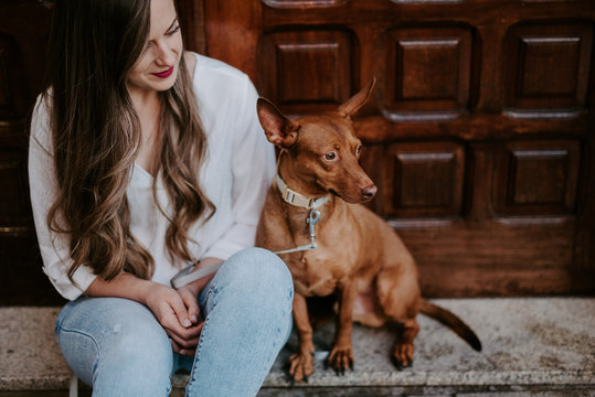 Beautiful casual woman with hound dog sitting on concrete step on street looking away