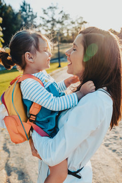 Young woman holding little girl with backpack while spending time in park together on sunny day
