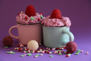arrangement aus zwei tassen gefüllt mit rosa creme mit himbeeren und streudekor obenauf, umgeben mit pralinen auf lila hintergrund