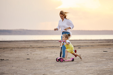Mother with kids riding scooters in summer. Woman runs along with her daughter on the beach at sunset. Daughter learns to ride a scooter © oes