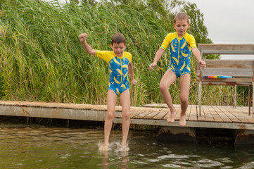Funny children in swimsuits accelerate on wooden bridge near river and jump into water. boys are happy on holiday in village together. Summer day, river, swimming in water
