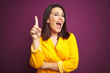 Young beautiful brunette woman wearing elegant yellow jacket over purple isolated background pointing finger up with successful idea. Exited and happy. Number one.