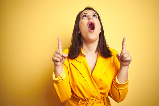 Young beautiful brunette woman wearing elegant dress over yellow isolated background amazed and surprised looking up and pointing with fingers and raised arms.
