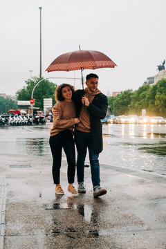 Side view of cheerful young man and woman with umbrella embracing while standing on wet city street on rainy day