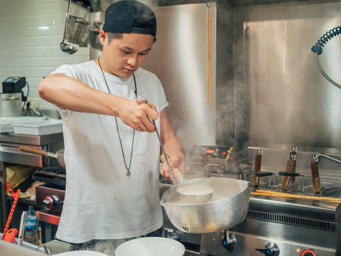 Young man in black cap pouring hot broth from saucepan for Japanese dish called ramen in Asian cafe