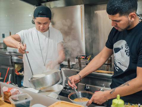 Young men pouring hot broth from saucepan for Japanese dish called ramen in Asian cafe