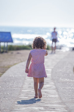 Back view of curly child in striped summer dress going along walkway on beach on blurred nature background