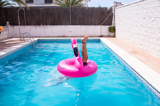 Young Fitness Man Jumping Into The Water In The Pool In The Center Of Inflatable Flamingo A Day Of Summer Vacation