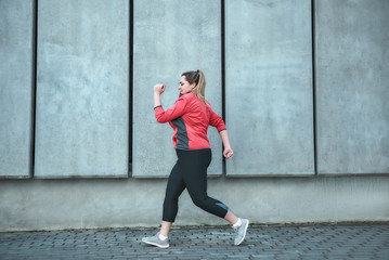 Morning workout. Side view of young plus size woman in sport clothes running outdoors.