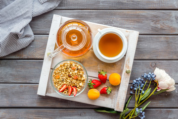 Breakfast in bed with granola, tea in pot and cup on tray on wooden background top view