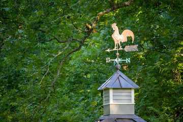 Close-up of a weathered copper rooster weathervane mounted on a wooden cupola with a weathered...