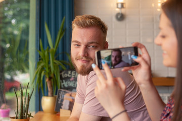 One girl talking a picture of a young man, on a smartphone. In a cafe.
