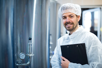 Technologist in white protective uniform and hairnet standing by chrome reservoirs with pressure gauge in food processing plant. Smiling industrial worker holding checklist. Quality control.